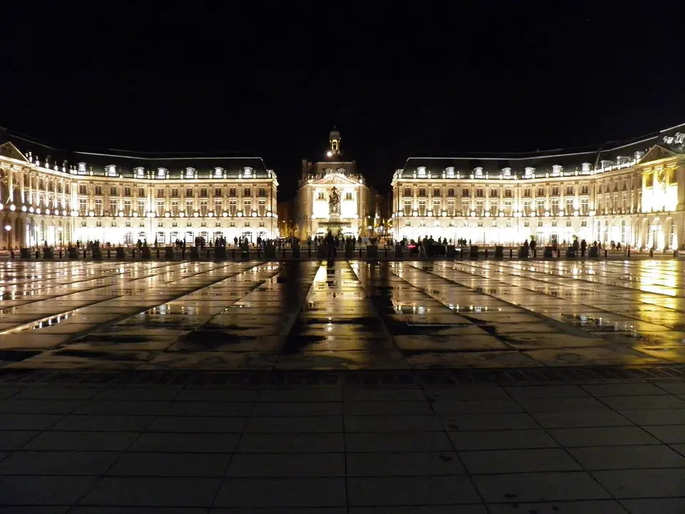 Place de la Bourse à Bordeaux de nuit, façades classiques en pierre éclairées et reflet des bâtiments sur le miroir d'eau.