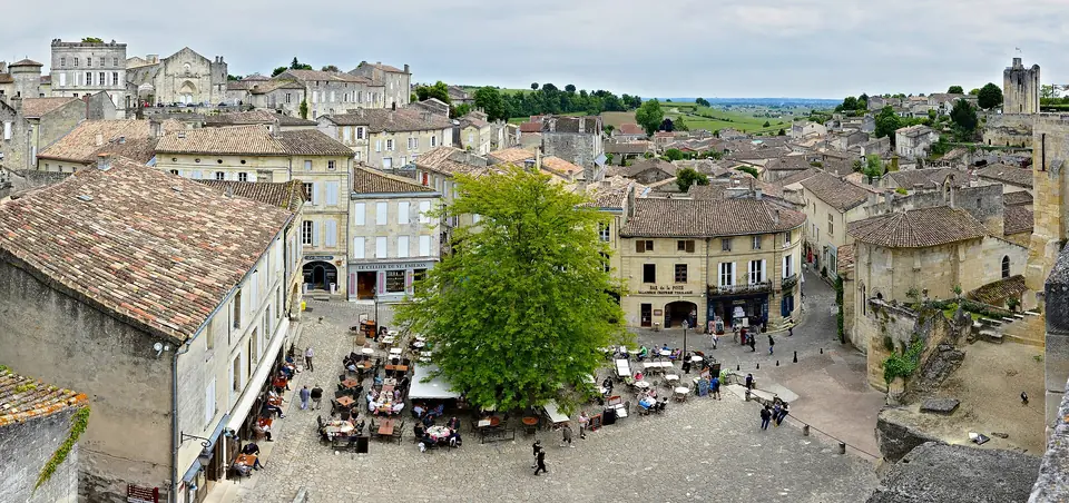 Vue plongeante sur la place principale de Saint-Émilion, ses terrasses de café et ses maisons en pierre blonde coiffées de tuiles.